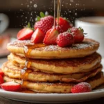 A stack of French toast pancakes topped with fresh strawberries and drizzled with maple syrup, served on a rustic wooden table with a coffee cup in the background.